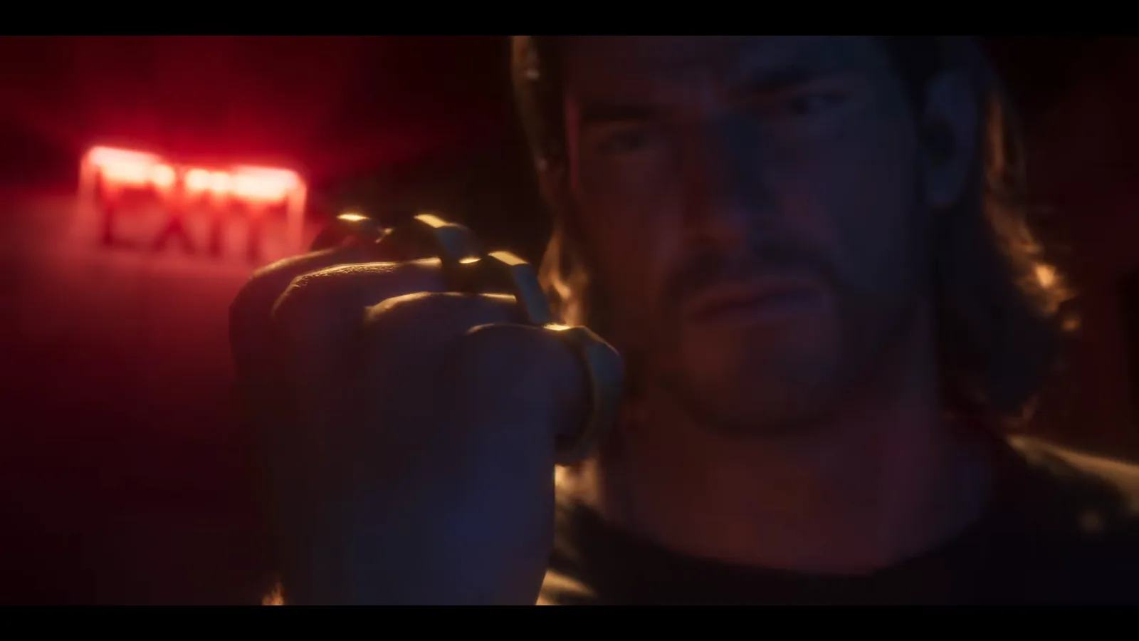 GTA 6 Launch Trailer: a long-haired man raises a clenched fist wearing gold knuckle dusters in front of a red EXIT sign